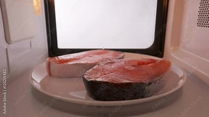 Using microwave oven to defrost salmon steaks before cooking it. Two fresh frozen fish steak on a plate spinning inside the microwave. Version without external lighting for more natural look.