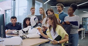 Multi-ethnic group of people surrounding robot. Humanoid working at computer. Colleagues noting everything on tablets and notepads. Woman giving commands. Testing hardware and operating system.