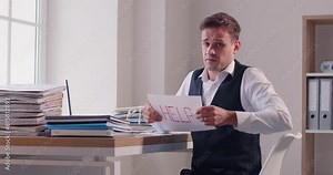 Bookkeeper asking for help with loads of paperwork. Tired man, sitting at office work desk with piles of paper, shows HELP sign and looks at camera with funny pleading imploring facial expression