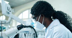 Close up of busy African American ауьфду medical researcher in white coat sitting at hospital laboratory using microscope, scientist looking at blood cells through equipment, experiment concept