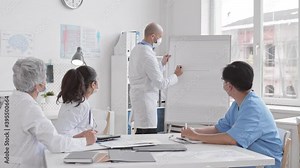 Hairless mature Caucasian doctor standing by flip chart board drawing system of axes, making presentation to diverse unrecognizable colleagues sitting at table in medical office