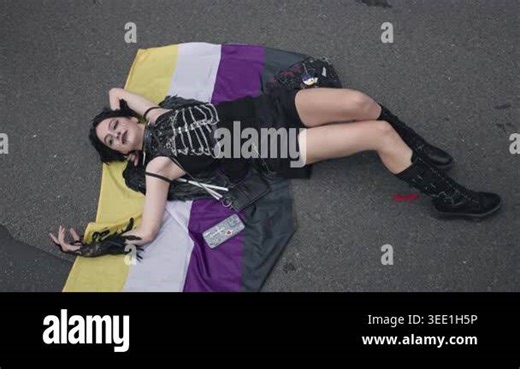 Berlin, Germany - 26.07.2025: A smiling person in black gothic attire with feather accessories lies on a nonbinary pride flag on the street during the Christopher Street Day parade Stock Video Footage - Alamy