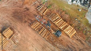 Professional machinery loads wooden trunks on stack putting in rows in plant yard. Heavy loader operates at rural sawmill near forest aerial view