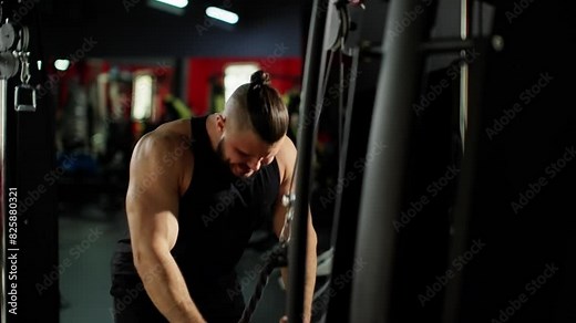 Muscular man in black tank top working out intensely on gym equipment, showing determination and strength, with a focus on upper body exercise