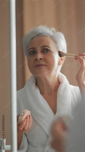 Rejuvenating lifting makeup for elderly lady, senior woman applying mineral powder on face skin. Vertical portrait of pretty aged woman looking at mirror in beauty routine, better aging, anti-aging