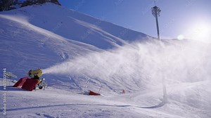 Snow cannon at ski resort in winter, snow machine produced artificial snow in mountains on slope at sunny day. Preparation of ski track in hills for active sport. Turbine spray water in cold weather.
