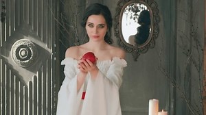 Fantasy queen beautiful woman evil witch holding red poison apple in hand offers to taste. Fairy girl gestures shows enchanted fruit. Vintage white dress. Princess lady beauty face in black dark room.