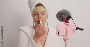 Women beauty blogging concept. Young female model does cosmetic makeup tutorial vlog poses at smartphone webcam applies hygienic lipstick cucumber mask wears bathrobe towel on head after taking shower