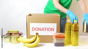 Food donation box. Girl packing humanitarian aid: canned goods, groceries, sunflower oil, and fruit in a cardboard box labeled Donation.