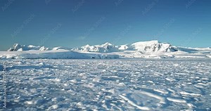 Fly over snowy mountain landscape in Antarctica. Polar frozen ocean landscape covered by snow under blue sunny sky. Discover the beauty of South Pole. Antarctica travel and exploration background