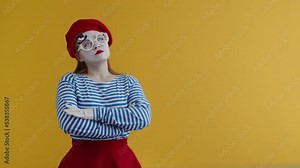Young mime woman in a suit and makeup looks up thoughtfully, holding her folded hands in front of her, on an orange background. Thinking, thinking