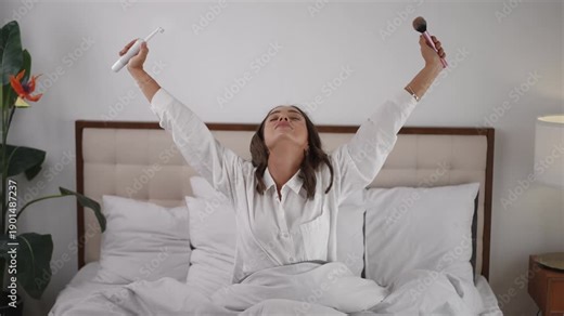 Happy young woman waking up in her bed, singing and dancing using an electric toothbrush and makeup brush. Carefree morning routine concept