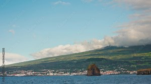 Approaching to Pico island by boat in the Azores, Portugal. View of the rocky coastline and local town. Pico mountain in the distance. Mount Peak covered with clouds.