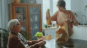 Young woman unloading groceries from paper bag on kitchen table and telling something to senior grandfather when taking care of him and helping with daily living at home