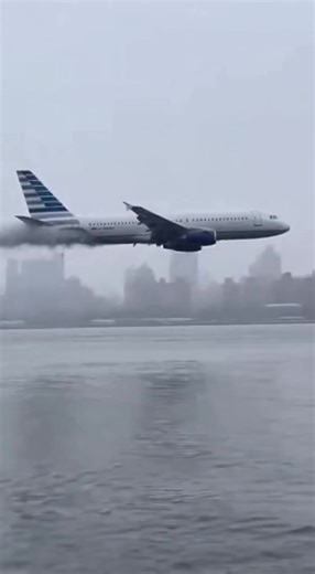 Ultra-realistic raw eyewitness footage of a commercial passenger jet flying dangerously low over a river near New York City after apparent engine failure. One engine trailing thick smoke as the aircraft struggles to maintain altitude. The jet clips the water surface before crashing fully into the river, creating massive spray and waves. Cold daylight, gray sky, distant skyline barely visible through haze. Slight handheld shake, imperfect framing. No cinematic effects, no color grading, no soundt