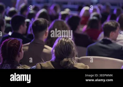 Audience seated in conference hall listening to speaker, purple stage lighting creates focused, contemplative atmosphere. Back view of diverse crowd in rows of seats Stock Video Footage - Alamy