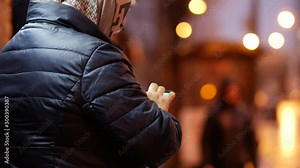 An elderly woman with glasses and an autumn dark quilted jacket types a message on the smartphone screen. Late evening. In the background a lot of night city lights. Touchscreen