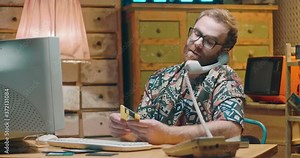 Caucasian young cheerful man sitting ar vintage PC monitor, holding diskette and talking on telephone. Retro style of interior. Male hacker or technician from 70's 80's at computer.