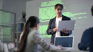 Medium footage of team of young programmers working on laptops listening to male tutor holding laptop explaining program coding using screen projector for demonstration