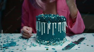 Pink hair birthday girl sitting at the table, taking piece of birthday cake and eating it with her hands