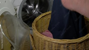 Closeup of a man's hands removing a tissue from a pocket of a pair of joggers or sweatpants, from a wicker laundry basket, before throwing the garment into an open washing machine.