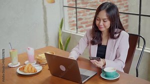 An Asian woman in a pink blazer is seated at a wooden desk, holding a credit card while making an online transaction on her laptop. A croissant, toast, and drinks are on the table in a cozy setting