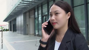 Asian female secretary is talking on the phone with a customer in front of her office building while traveling.