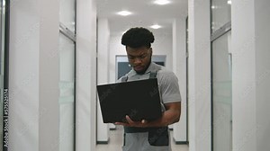 African American installer in uniform stands in corridor and sets up security cameras using software on laptop. CCTV camera installation in business office. Concept of surveillance system. Dolly zoom.
