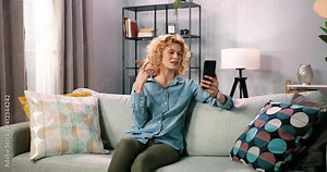 Happy girl sitting on sofa at home, holding smartphone and looking at screen while having video call at distance with friend. Online mobile chat app using smartphone. Videochat application concept