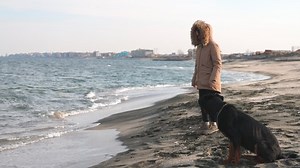 An attentive, kind, demanding hostess in a jacket trains and feeds her large well-mannered faithful dog of the Rottweiler breed on a sandy beach near the sea, 4K UHD slow-motion video