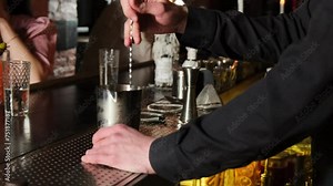 Experienced barkeeper mixes alcoholic beverage with spoon in bar interior. Bar equipment surrounds aluminum glass standing on counter