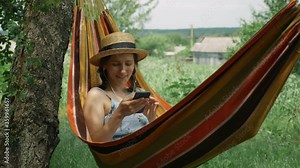 Woman in earphones lying in hammock and listening to music, singing and relaxing at green garden. Cute smiling girl in hat and dress resting in hammock and listens to music from earphones