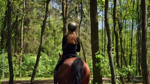 Live camera follows unrecognizable woman riding horse in summer spring forest on sunny day. Back view confident Caucasian young slim equestrian on back of graceful animal walking in sunshine