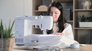 The beautiful young girl sits at the automatic sewing machine in sewing studio and sews some clothes, the brunette, long hair, flowers and accessories