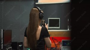 Elegant woman singer with headphones in black dress with open back performs song with microphone dancing in recording studio close backside view