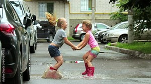 Two little girls in pink rubber boots are joyfully jumping in a rain puddle after a rainstorm in the courtyard of a high-rise building among cars. Slow motion. 4k video.