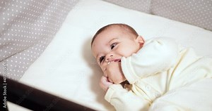 Newborn cheerful littlebaby is chewing and playing in his crib with nipple. Funny smiling looks through the railing of the crib. slow motion