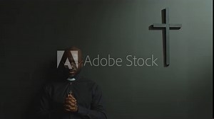 Black Catholic priest wearing cassock standing against grey wall with minimalist cross, holding hands clasped and eyes closed as praying to God in modern church