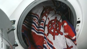 Person's hand puts a towel inside the steel drum of the washing machine. White washing machine with front-loading laundry, door of laundry load with clear glass for monitoring operation. Close-up