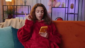 Preteen girl texting share messages content on smartphone social media applications online watching relax movie, news. Young child, kid at home in night evening living play room sitting on orange sofa