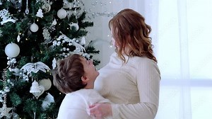 Joyful mother lifts and hugs her laughing son near a beautifully decorated christmas tree. Both wear white outfits, radiating happiness and warmth in a festive home setting