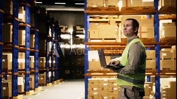 Male personnel operating with laptop in e-commerce distribution center, counting parcels and merchandise on pallets. Staff ensures supply chain shipment and package tracking operations.