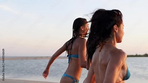 Group of happy wet girls in bikini running into sea splashing water on sunset. Cheerful female friends having fun on beach near pond. Young women go to swim and bathe in lake. Slow motion splashes.
