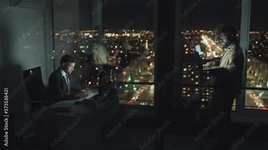 Full shot of two male Caucasian and black analysts working at night in dark office, sitting at desk typing on computer, and standing with laptop, filmed through window, with reflections on glass