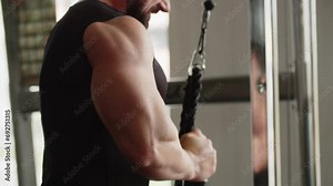 A young man bodybuilder in the gym trains his arms muscles on a exercise machine and demonstrates power of them. Bending and extending arm up and down. Lifting of cables with weights. A man is