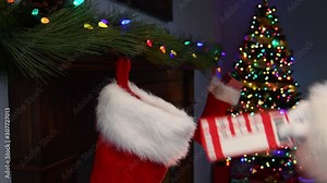 A close-up of Santa Claus’s hand placing a pair of blank ticket stubs into a Christmas Stocking hanging on a mantelpiece with a Christmas tree with twinkling lights in the background Stock Video