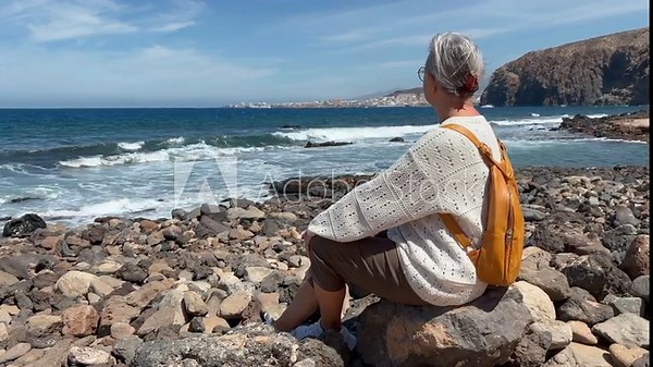 Rear view of senior grey haired woman sitting at the rocky beach looking the waves crashing on the rocks in a windy day. Elderly woman in retirement or vacation enjoying sunny day and freedom