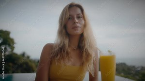 A blonde woman drinks mango shake while lounging in the sun by the pool on vacation. Elegant young woman drinks cocktail by the pool in slow motion.