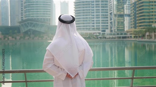 Emirati man wearing traditional white clothing standing at the Dubai Marina promenade and watching the canal, rear view.