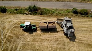 Aerial view gray combine harvester with full hopper auger unloading pours wheat or barley in trailer with tractor on agricultural field near road. Drone shoots video of reap grain crops
